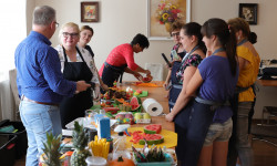 w sali przy długim stole na którym leża produkty spożywcze kilka kobiet i mężczyzna jedna z kobiet kroi pozostałe osoby z sobą rozmawiają