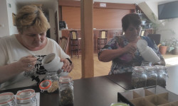 dwie kobiety przy stole nakładają z pojemników łyżką susz ziołowy do szklanych małe słoiki 