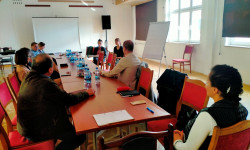 grupa uczestników spotkania siedzących przy stole słuchają wypowiedzi osoby prowadzącej